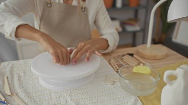 Kadın bej bir önlük giyerken bir çömlek atölyesinde çıplak elleriyle kil yoğurur; sakin odaklı yaratıcılık.