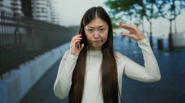 Şehir sokağında telefonla dalga geçen, günlük kıyafetler giyen, uzun saçlı, şehir yolunda esprili bir şekilde el kol hareketi yapan, oyuncu ifadeler sergileyen, açık hava sahnesi olan bir kadın..