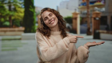Genç bir kadın, açık hava parkında uzanan avuç içini göstererek gülümsüyor, rahat bir kazak giyiyor, güneşli günün tadını çıkarıyor ve bulanık yeşillik ve kentsel unsurlarla çevrili..