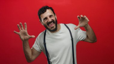 Sakallı, beyaz gömlek ve pantolon askısı giymiş, neşeli ve enerjik bir hava saçan, neşeli kırmızı bir arka planda poz veren İspanyol bir adam..