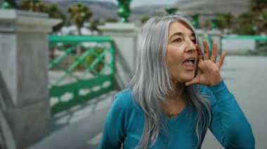 Gri saçlı ve mavi gömlekli bir kadın sokakta bağırıyor, yeşil ve bulanık arka planlı açık şehir manzarası canlı bir ortam olduğunu gösteriyor..