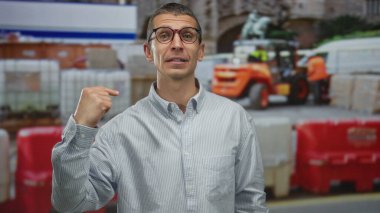 Gözlüklü beyaz adam, arka planda inşaat işçileri ve araçlarla dolu işlek bir caddede kendini işaret ediyor..