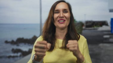 Sahilde sevinçle kutlama yapan bir kadın, plaj zemininde ışıl ışıl gülümserken, dışarıda gülümseyen genç İspanyol bayanın neşeli havasını vurguluyor..