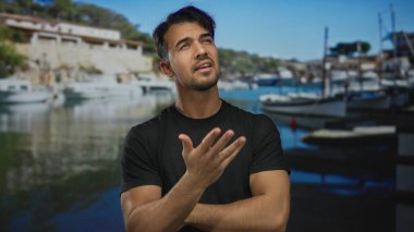 Düşünceli bir suratı olan genç İspanyol bir adam tekneleri ve sakin bir açık hava ortamı olan manzaralı bir sahil limanında duruyor..