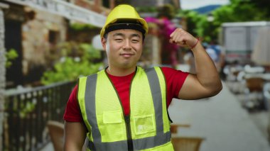 İnşaat malzemesi giymiş bir adam sokak ortasında kaslarını esnetiyor, sarı bir şapka ve yansıtıcı yelek giyerken gücünü ve güvenini gösteriyor..