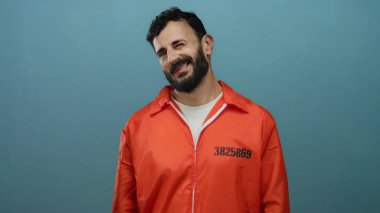 Turuncu hapishane tulumu giyen İspanyol sakallı adam mavi bir arka plana gülümsüyor ve samimi ve rahat bir ifade sergiliyor..