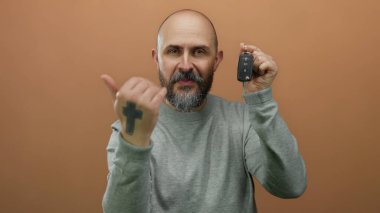 Sakallı bir adam araba anahtarlarını kahverengi duvara doğrultuyor. Otomotiv ya da yaşam tarzı içeriği için jest ve ifade sergiliyor..