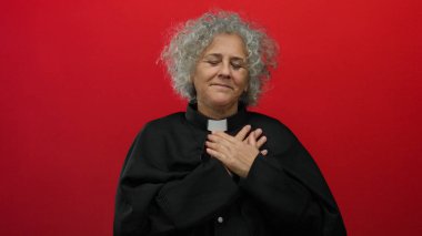 Gri saçlı, ruhban kıyafeti giyen ve göğsünün üstünde canlı kırmızı izole edilmiş arka planda el ele tutuşan barış ve ruhaniliği ifade eden bir kadın..