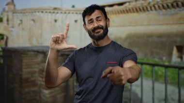 Roma 'da antik mimarinin önünde şakacı bir jest yapan sakallı İspanyol bir adam, tarihi bir taş arka plana işaret ediyor ve gülümsüyor..