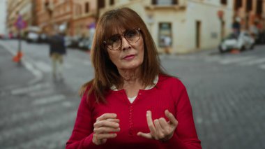 Kırmızı kazaklı yaşlı İspanyol kadın hareketleriyle kalabalık bir şehir caddesinde canlı şehir yaşamını resmediyor..