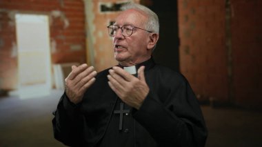 Siyah cüppeli kıdemli rahip, duvarlarla örülü bir inşaat alanında düşünceli bir şekilde el kol hareketi yapar, kırsal bir alanda tefekkür ve kendini adama anını yakalar..