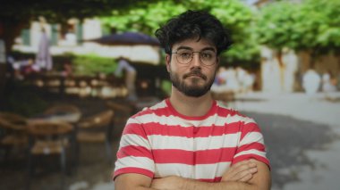 Gözlük takan sakallı genç adam güvenli bir şekilde ayakta duruyor. Arka planı bulanık, hareketli ve benzersiz bir sokak manzarası ve eşsiz bir stili olan hareketli bir sokakta kesişiyor..