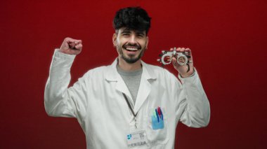 Kırmızı duvarın önünde elinde optik aletle gülümseyen beyaz önlüklü bir adam mutluluk ve profesyonellik gösteriyor..
