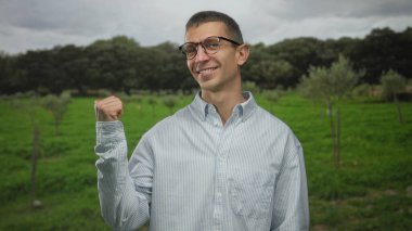 Canlı bir açık hava parkı ortamını işaret eden, gözlüklü ve çizgili tişörtlü bir adam bulutlu gökyüzünün altında neşeli ve kendinden emin bir ifade sergiliyor..