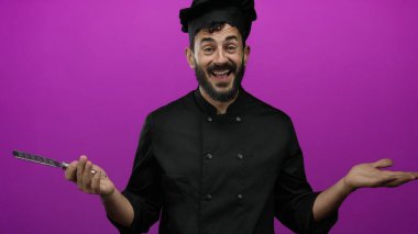 Siyah üniformalı İspanyol şef elinde peynir rendesiyle canlı pembe arka planda sırıtıyor..