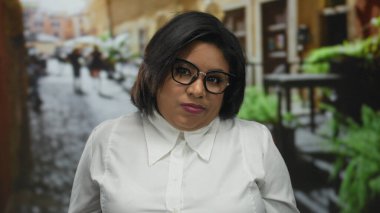 Şehir caddesinde gözlüklü bir kadın, kısa saçlı ve beyaz gömlekli, bulanık kentsel bir çevreyle çevrili kendinden emin Latin görünümünü sergiliyor..