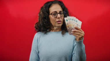 Orta yaşlı bir kadın, kırmızı bir duvara karşı Arjantin banknotları tutuyor. Finansal stres ve tefekkür duygularını aktarıyor..