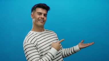 Çizgili gömlekli genç adam gülümsüyor ve mavi arka plana işaret ediyor.
