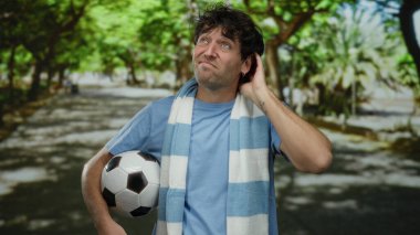 Parkta futbol oynayan İspanyol bir adam, güneşli yeşil bir yolda dikilirken belirsizliğini ifade eder..