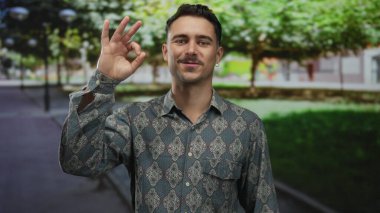 Genç İspanyol adam desenli gömleğiyle gülümsüyor, sakin parkta işaret veriyor yemyeşil ağaçların altında ve sessiz arka planda, rahat açık hava atmosferini yakalıyor..