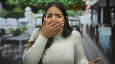 Beyaz kazaklı Latin bir kadın, yeşil ağaçlar ve bulanık yaya geçmişi ile çevrili bir restoranın terasında şaşırmış bir ifade sergiliyor..