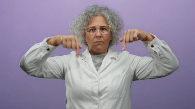 Beyaz önlüklü kadın mor arka planda aşağı doğru hareketler yapıyor, kendinden emin orta yaşlı bir yüz ifadesi sergiliyor..