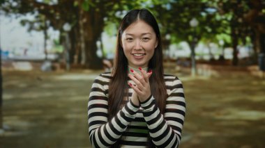 Açık havada gülümseyen bir kadın, güneşli bir parkta yeşil ağaçlı çizgili bir süveter giyiyor, neşeli bir ifade ve doğal güzellik sergiliyor canlı, huzurlu bir açık hava ortamında..