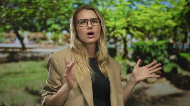 Yeşil bir parkta tutkulu bir şekilde konuşan, gözlüklü ve bej ceketli, gün boyunca yemyeşil, hareketli jestler yapan bir kadın..
