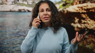 Sahil kenarında kıvırcık saçlı, bulanık arka planı ve berrak mavi suyu olan, sakin bir açık hava sahili tasvir eden bir kadın telefonda konuşuyor..