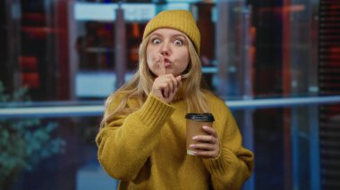 Kadın, modern bir otel lobisinde elinde kahve tutarken, sarı kazak ve bere giyerken, ciddi ifadesini çerçeveleyen sarı saçlı, sessiz kalma ihtiyacını gösteren bir jest yapıyor.