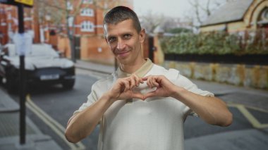 Sokaklarda dikilen bir adam elleriyle kalp şekli yaratıyor, binalar ve park halindeki arabalarla kentsel çevre tarafından geliştirilmiş sıradan bir ortamda mutluluk ve pozitiflik yayıyor..