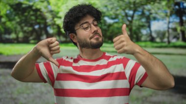 Çizgili tişörtlü, sakallı genç adam yeşil bir parkta karışık duygular ve duyguları yansıtan jestler yapıyor..