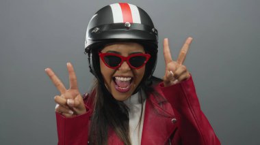Kasklı ve kırmızı ceketli bir kadın, izole edilmiş gri arka planda barış işareti ile poz veriyor, gençlik enerjisi ve kendine güveni yayıyor..