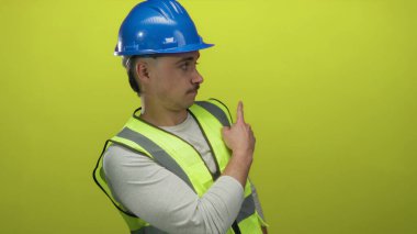 Yansıtıcı yelek giyen ve kasketi parmağıyla izole edilmiş sarı arka plan duvarını işaret eden İspanyol genç adam, güvenlik odaklı bir inşaat konseptini gösteriyor..