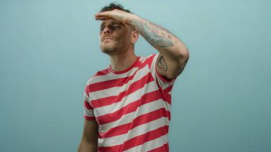 İspanyol erkek dikkatle uzaklığa bakıyor, saf mavi bir arka plana karşı duruyor, dövmelerini ve kendinden emin duruşu olan günlük çizgili gömleğini vurguluyor..