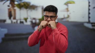 Kırmızı tişörtlü genç adam güneşli bir şehirde beyaz binalar ve palmiye ağaçlarıyla çevrili bir sokakta yumruklarıyla şakacı pozlar veriyor..