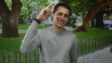 Arka planda ağaçlarla dolu bir parkta gülümseyen genç adam, sıradan bir görünüm sergiliyor..