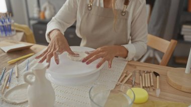 Genç sarışın bir kadın, stüdyoda çıplak elle odun bir masanın üzerinde çıplak elle yoğurulan bir önlük giyiyor; yaratıcılık odaklı sanatçılık.