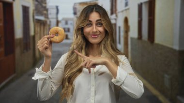 Genç sarışın kadın elinde tuttuğu donutu işaret ediyor güneş ışığında kaldırım taşı caddesinde; neşe aşeriyor.