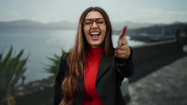 Gözlüklü gülümseyen bir kadın, güneşli deniz kıyısında arka planda dağlar olan kameraya doğru uzanır. Bu da sıcaklık ve açıklığı gösterir..