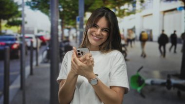 Şehir caddesinde insanlarla çevrili akıllı telefon kullanırken gülümseyen bir kadın ve güneşli bir günde açık havada günlük giysiler giymiş, odaklanmış ve rahat görünen bir kadın..