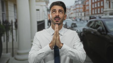 Beyaz gömlekli ve lacivert kravatlı genç bir adam park halindeki arabalarla dolu bir caddede çıplak ellerini birbirine bastırıyor..