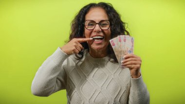Uae 'den orta yaşlı bir kadın dişlerini gösterirken izole edilmiş sarı bir zemin üzerinde Dirham banknotları tutuyor. Finansal başarı ve diş sağlığı gösteriyor..