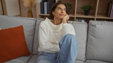 Kadın rahat bir oturma odasında koltukta dinleniyor, rahat bir kıyafet giyiyor, düşünceli görünüyor, arka planda raflar ve bitkiler var, sıcak bir ev havası yaratıyor..