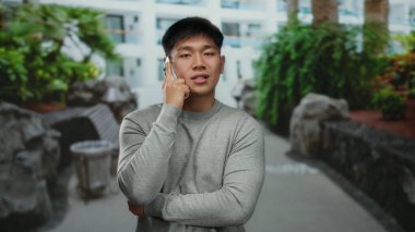 Genç adam, yeşil bitkiler ve modern binalarla dolu bir caddede yürürken akıllı bir telefonla konuşuyor, dinamik bir şehir manzarasında şehir yaşamını gözler önüne seriyor..