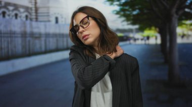 Şehrin dışındaki genç İspanyol kadın gözlük ve ceket takıyor. Ağaçlarla ve arka planda bulanık binalarla düşünceli görünüyor..