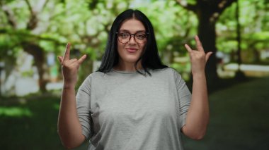 Parkta gözlüklü bir kadın güneşli bir günde gülümseyen gri tişörtlü, güveni ve güveni olan ağaçlarla dışarıda taş jestler yapıyor..