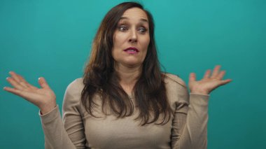 Uzun saçlı bir kadın yeşil arka plan duvarının üzerinden omuz silkip, bej bir kazak giyiyor ve kafa karışıklığını dile getiriyor..