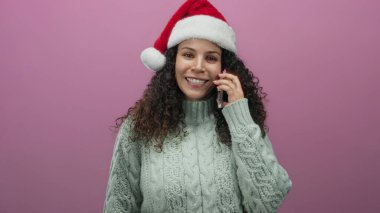 Noel Baba şapkalı genç kadın telefonda pembe arka planda konuşuyor, rahat bir kazak giyiyor ve bayram boyunca mutlu bir şekilde gülümsüyor..