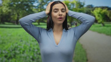 Parkta elleri kafasında, mavi gömlekli, yeşil doğayla çevrili bir kadın, güneşli bir günde bulanık arka planda duygularını gösterir..